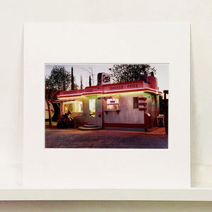 Mounted photograph by Richard Heeps. This photograph depicts a one storey small building "Dot's Diner" brightly lit with a pink roof, with Hamburgers, Hot Dogs, Shakes, Fries written along the top width of the building.