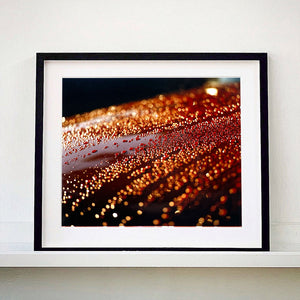 Black framed photograph by Richard Heeps. The photograph is of a brown metal surface with water droplets on. The lighting makes the droplets appear in different colours of orange and brown.