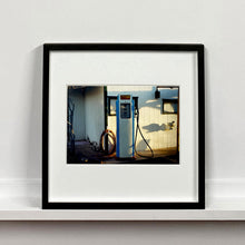 Load image into Gallery viewer, Black framed photograph by Richard Heeps. A vintage petrol pump with a white front and blue sides, sitting outside a white slatted building.
