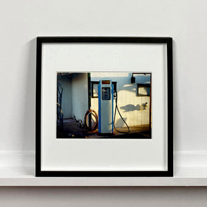 Black framed photograph by Richard Heeps. A vintage petrol pump with a white front and blue sides, sitting outside a white slatted building.