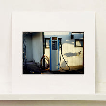 Load image into Gallery viewer, Mounted photograph by Richard Heeps. A vintage petrol pump with a white front and blue sides, sitting outside a white slatted building.