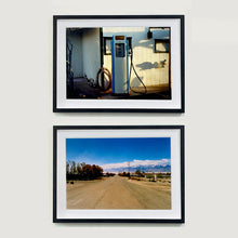 Load image into Gallery viewer, Two black framed photographs held by Richard Heeps. The top photograph depicts a vintage petrol pump with a white front and blue sides, sitting outside a white slatted building.  The bottom photograph shows a dusty road in the middle, heading towards the snow capped mountains in the distance, on the right are brown bushes and trees and on the left, single level concrete buildings. 