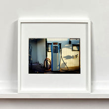 Load image into Gallery viewer, White framed photograph by Richard Heeps. A vintage petrol pump with a white front and blue sides, sitting outside a white slatted building.