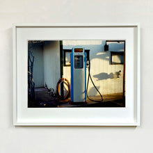 Load image into Gallery viewer, White framed photograph by Richard Heeps. A vintage petrol pump with a white front and blue sides, sitting outside a white slatted building.