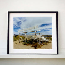 Load image into Gallery viewer, Black framed photograph by Richard Heeps. The back view of a basic white painted grandstand sits in the middle of this photograph. Grass grows up from its base and it sits alone on a sandy ground.