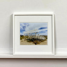 Load image into Gallery viewer, White framed photograph by Richard Heeps. The back view of a basic white painted grandstand sits in the middle of this photograph. Grass grows up from its base and it sits alone on a sandy ground.