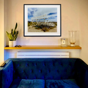 In situ photograph by Richard Heeps. The back view of a basic white painted grandstand sits in the middle of this photograph. Grass grows up from its base and it sits alone on a sandy ground.