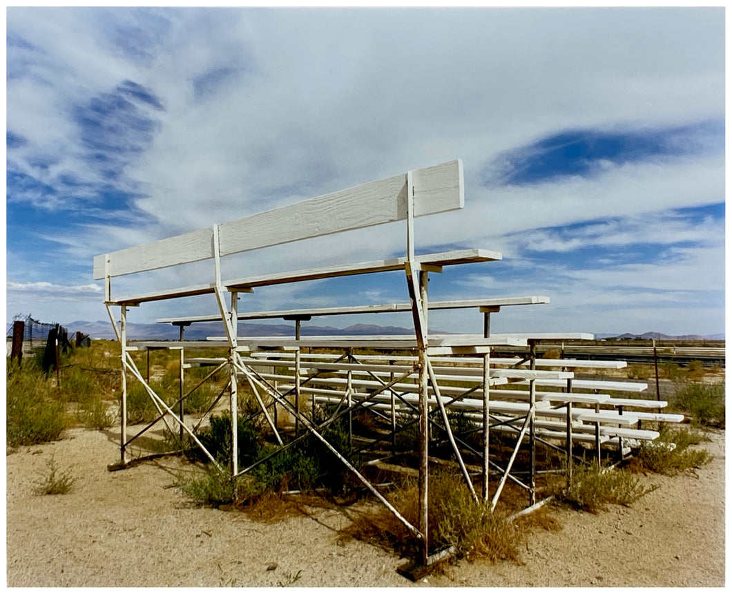 Photograph by Richard Heeps. The back view of a basic white painted grandstand sits in the middle of this photograph. Grass grows up from its base and it sits alone on a sandy ground.