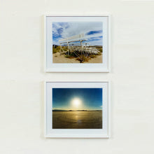 Load image into Gallery viewer, Two white framed photographs by Richard Heeps. The back view of a basic white painted grandstand sits in the middle of the top photograph. Grass grows up from its base and it sits alone on a sandy ground. The bottom photograph depicts sand with circles in, hills in the distance and a blue sky glowing with a hazy sun in the middle.