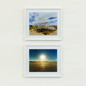 Two white framed photographs by Richard Heeps. The back view of a basic white painted grandstand sits in the middle of the top photograph. Grass grows up from its base and it sits alone on a sandy ground. The bottom photograph depicts sand with circles in, hills in the distance and a blue sky glowing with a hazy sun in the middle.