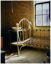 Load image into Gallery viewer, Photograph by Richard Heeps. A run down now unused room with the metal surround of a cot bed and no mattress, at the end of the bed a wash stand with a bowl on top. Light shines in the room from the window.