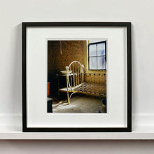 Load image into Gallery viewer, Black framed photograph by Richard Heeps. A run down now unused room with the metal surround of a cot bed and no mattress, at the end of the bed a wash stand with a bowl on top. Light shines in the room from the window.