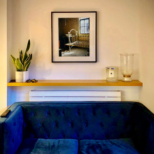 In situ photograph by Richard Heeps. A run down now unused room with the metal surround of a cot bed and no mattress, at the end of the bed a wash stand with a bowl on top. Light shines in the room from the window.