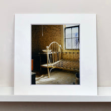 Load image into Gallery viewer, Mounted photograph by Richard Heeps. A run down now unused room with the metal surround of a cot bed and no mattress, at the end of the bed a wash stand with a bowl on top. Light shines in the room from the window.