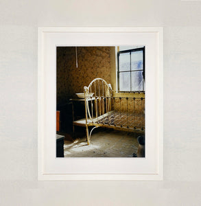 White framed photograph by Richard Heeps. A run down now unused room with the metal surround of a cot bed and no mattress, at the end of the bed a wash stand with a bowl on top. Light shines in the room from the window.