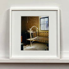 Load image into Gallery viewer, White framed photograph by Richard Heeps. A run down now unused room with the metal surround of a cot bed and no mattress, at the end of the bed a wash stand with a bowl on top. Light shines in the room from the window.