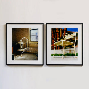 Two black framed photographs by Richard Heeps. The photograph on left hand side is of a run down now unused room with the metal surround of a cot bed and no mattress, at the end of the bed a wash stand with a bowl on top. Light shines in the room from the window. The photograph on the right hand side is a metal chair sitting with an out of focus background which has trees, blue sky and grass.