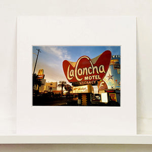Mounted photograph by Richard Heeps. This photograph is of the outside of La Concha Motel. The gold flamboyant La Concha lettering is set on a big red background. Below the motel sign is NO VACANCY with just VACANCY lit in red, below this sits a sign for Budget rent a car. Other signs and palm trees are the background together with a blue sky.