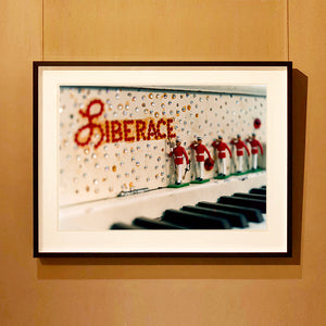 Black framed photograph by Richard Heeps. A close up of Liberace's piano. This captures the black keys and the fall board which is decorated with crystals and red buttons spelling out Liberace in capitals. There are also 5 red and white metal soldiers positioned marching along the rim.