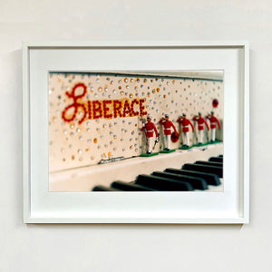 White framed photograph by Richard Heeps. A close up of Liberace's piano. This captures the black keys and the fall board which is decorated with crystals and red buttons spelling out Liberace in capitals. There are also 5 red and white metal soldiers positioned marching along the rim.
