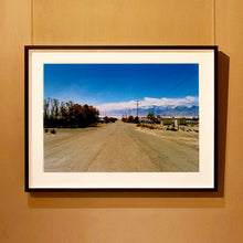 Load image into Gallery viewer, Black framed photograph by Richard Heeps. A dusty road in the middle, heading towards the snow capped mountains in the distance, on the right are brown bushes and trees and on the left, single level concrete buildings.