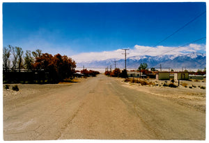 Photograph by Richard Heeps. A dusty road in the middle, heading towards the snow capped mountains in the distance, on the right are brown bushes and trees and on the left, single level concrete buildings.