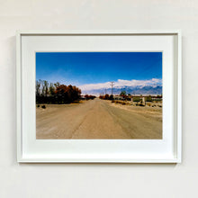 Load image into Gallery viewer, White framed photograph by Richard Heeps. A dusty road in the middle, heading towards the snow capped mountains in the distance, on the right are brown bushes and trees and on the left, single level concrete buildings.