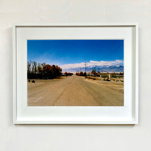 White framed photograph by Richard Heeps. A dusty road in the middle, heading towards the snow capped mountains in the distance, on the right are brown bushes and trees and on the left, single level concrete buildings.