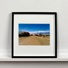 Load image into Gallery viewer, Black framed photograph by Richard Heeps. A dusty road in the middle, heading towards the snow capped mountains in the distance, on the right are brown bushes and trees and on the left, single level concrete buildings.