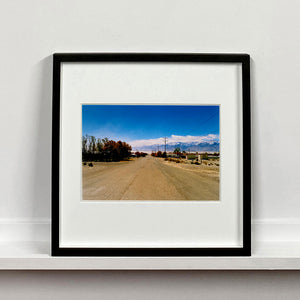 Black framed photograph by Richard Heeps. A dusty road in the middle, heading towards the snow capped mountains in the distance, on the right are brown bushes and trees and on the left, single level concrete buildings.