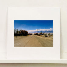 Load image into Gallery viewer, Mounted photograph by Richard Heeps. A dusty road in the middle, heading towards the snow capped mountains in the distance, on the right are brown bushes and trees and on the left, single level concrete buildings.