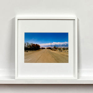 White framed photograph by Richard Heeps. A dusty road in the middle, heading towards the snow capped mountains in the distance, on the right are brown bushes and trees and on the left, single level concrete buildings.