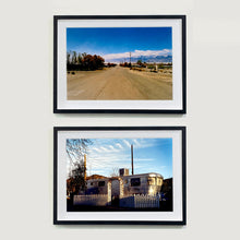 Load image into Gallery viewer, Two black framed photographs by Richard Heeps. The top photograph shows a dusty road in the middle, heading towards the snow capped mountains in the distance, on the right are brown bushes and trees and on the left, single level concrete buildings. The bottom photograph has a rounded metal caravan surrounded by a white picket fence with an archway entrance.