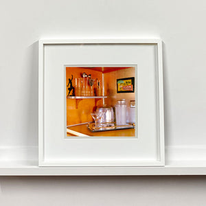 White framed photograph by Richard Heeps. A drinks corner with martini glasses, cocktail shaker and ice bucket. There is a shelf with cocktail making equipment and it is set against retro orange walls.