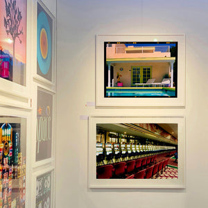 At the Art show - photographs by Richard Heeps. The top photograph shows Palm Springs Pool Side and showcases classic mid-century Palm Springs California architecture. Cool blue skies and pool with accents of pink and almost neon yellow. The bottom photograph features a row of slot machines and two rows of red chairs in front.