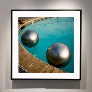 Black framed photograph by Richard Heeps. The corner of a circular swimming pool with two metallic silver beach balls floating on the water.