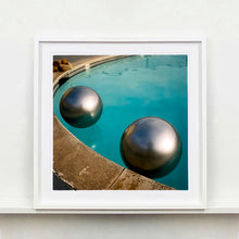 Load image into Gallery viewer, White framed photograph by Richard Heeps. The corner of a circular swimming pool with two metallic silver beach balls floating on the water.