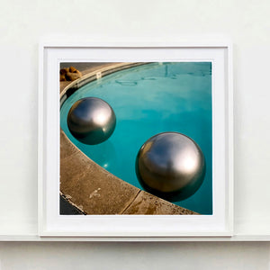 White framed photograph by Richard Heeps. The corner of a circular swimming pool with two metallic silver beach balls floating on the water.