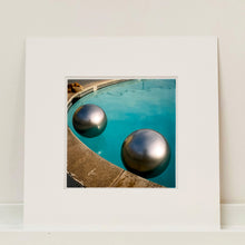 Load image into Gallery viewer, Mounted photograph by Richard Heeps. The corner of a circular swimming pool with two metallic silver beach balls floating on the water.