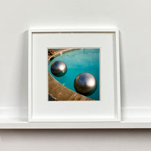 Load image into Gallery viewer, White framed photograph by Richard Heeps. The corner of a circular swimming pool with two metallic silver beach balls floating on the water.