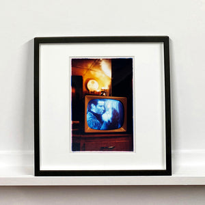 Black framed photograph by Richard Heeps. A retro small brown television sits on a wooden table displaying a black and white film which shows as blue and white. In the background of the television is a warm light.