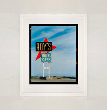 Load image into Gallery viewer, White framed photograph by Richard Heeps edged by film rebate. A roadside sign on Route 66 in America. The word ROY&#39;S appears in a black sign with a bit red arrow pointing to the left ground, below this VACANCY and on a green square the words MOTEL and CAFE.