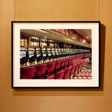 Load image into Gallery viewer, Black framed photograph by Richard Heeps.  The photograph depicts a line of slot machines sitting in a vintage Las Vegas casino. Perspective moves the slot machines from big machines on the left hand side to smaller on the right hand side. In front of the machines are a row of neatly lined blue stools one per machine and then red spectators chairs are neatly lined sitting forefront of the shot.