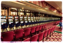 Load image into Gallery viewer, Photograph by Richard Heeps. A line of slot machines sits in a vintage Las Vegas casino. Perspective moves the slot machines from big machines on the left hand side to smaller on the right hand side. In front of the machines are a row of neatly lined blue stools one per machine and then red spectators chairs are neatly lined sitting forefront of the shot.