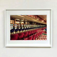 Load image into Gallery viewer, White framed photograph by Richard Heeps. A line of slot machines sits in a vintage Las Vegas casino. Perspective moves the slot machines from big machines on the left hand side to smaller on the right hand side. In front of the machines are a row of neatly lined blue stools one per machine and then red spectators chairs are neatly lined sitting forefront of the shot.