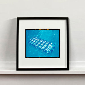 Black framed photograph by Richard Heeps. A transparent blue lilo is sun bathing on a blue pool. 