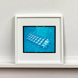 White framed photograph by Richard Heeps. A transparent blue lilo is sun bathing on a blue pool. 