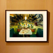 Load image into Gallery viewer, Black framed photograph by Richard Heeps. Inside a trailer, there is a fixed table and chairs with two tea cups and saucers on the table. Behind on the shelf is a tea set and a doll dressed as a bride.