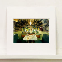 Load image into Gallery viewer, Mounted photograph by Richard Heeps. Inside a trailer, there is a fixed table and chairs with two tea cups and saucers on the table. Behind on the shelf is a tea set and a doll dressed as a bride.