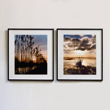 Load image into Gallery viewer, Two black framed photographs by Richard Heeps. On the left hand side is a photograph of reeds standing tall and reflecting down onto the water with a setting sun behind them. The photograph on the right hand side is a tussock sitting in the water with the dark clouds above reflecting into the surrounding water, bathed in an evening sun.